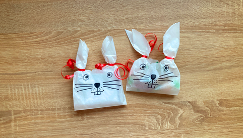 Zwei dekorierte Geschenktüten in Hasenform mit roten Schleifen und aufgemalten Gesichtern auf einem Holzuntergrund.