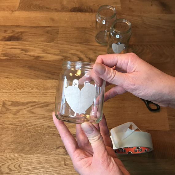 Hand, die ein beschriftetes Glas mit einem Herzmotiv hält, auf einem Holztisch, umgeben von weiteren leeren Gläsern und Klebeband.