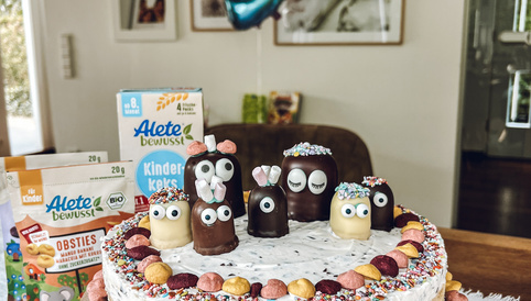 Bunter Geburtstagskuchen mit süßen Figuren, Streuseln und einer „9“-Luftballon. Verpackungen von Alete Produkten im Hintergrund.