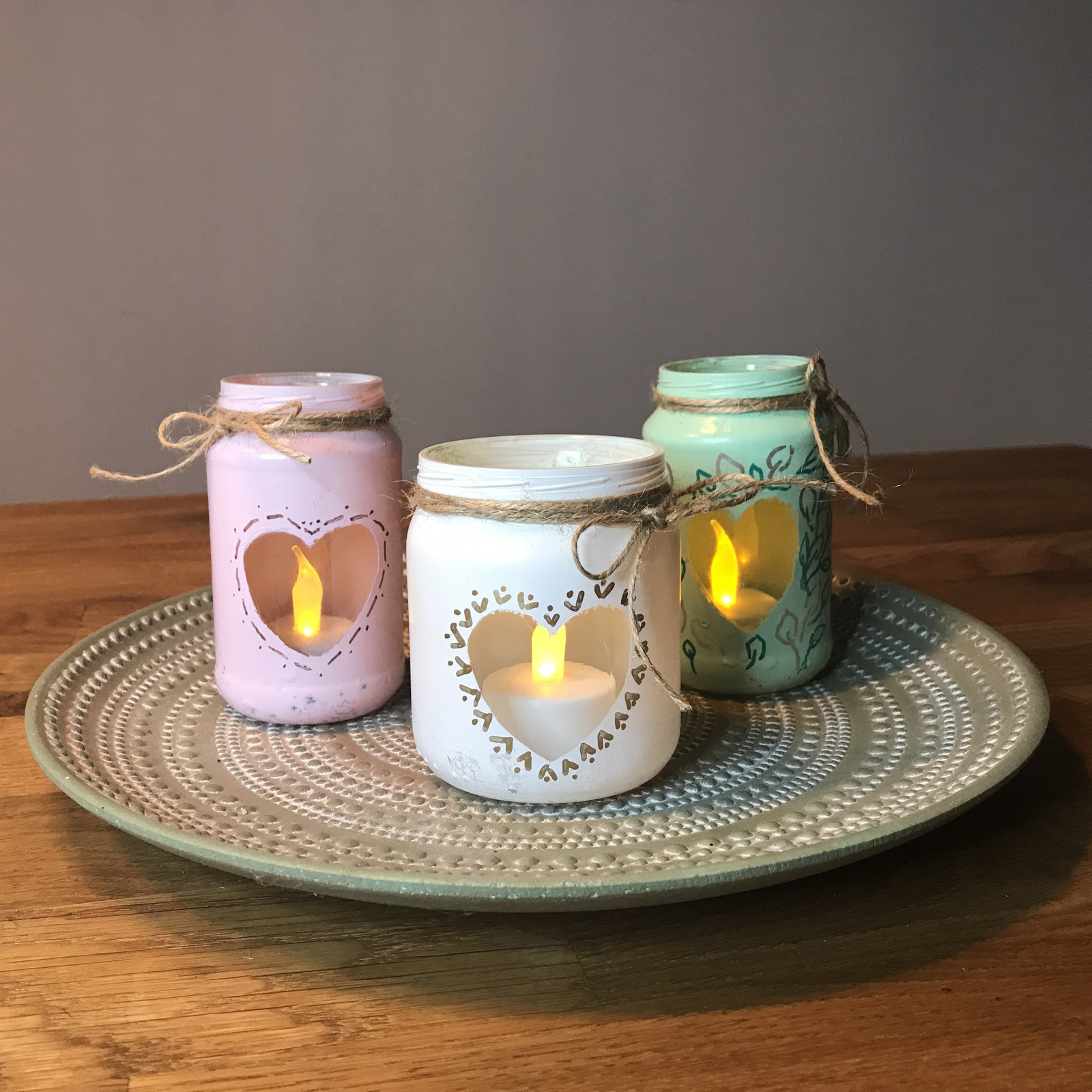 Dekorative Gläser mit Herzen, in pink, weiß und grün, auf einem stilvollen Teller, angezündet mit LED-Kerzen für gemütliche Atmosphäre.