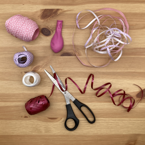 Bastelmaterialien auf einem Holztisch: Bänder in Pink- und Lilatönen, eine pinke Ballonform, Schere und Klebeband.