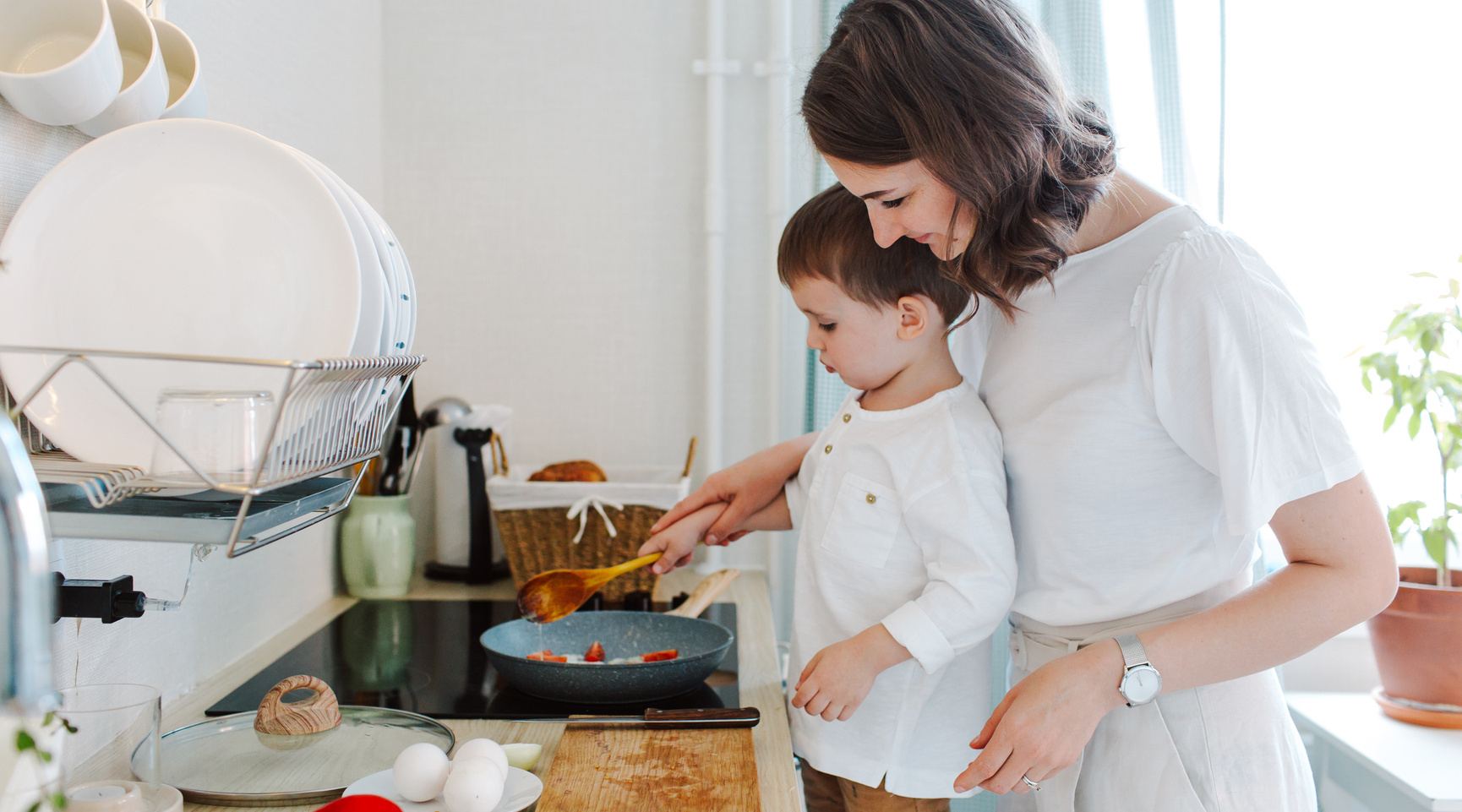 Junge hilft seiner Mutter beim Kochen in einer modernen Küche, während sie gemeinsam Zutaten in einer Pfanne zubereiten.