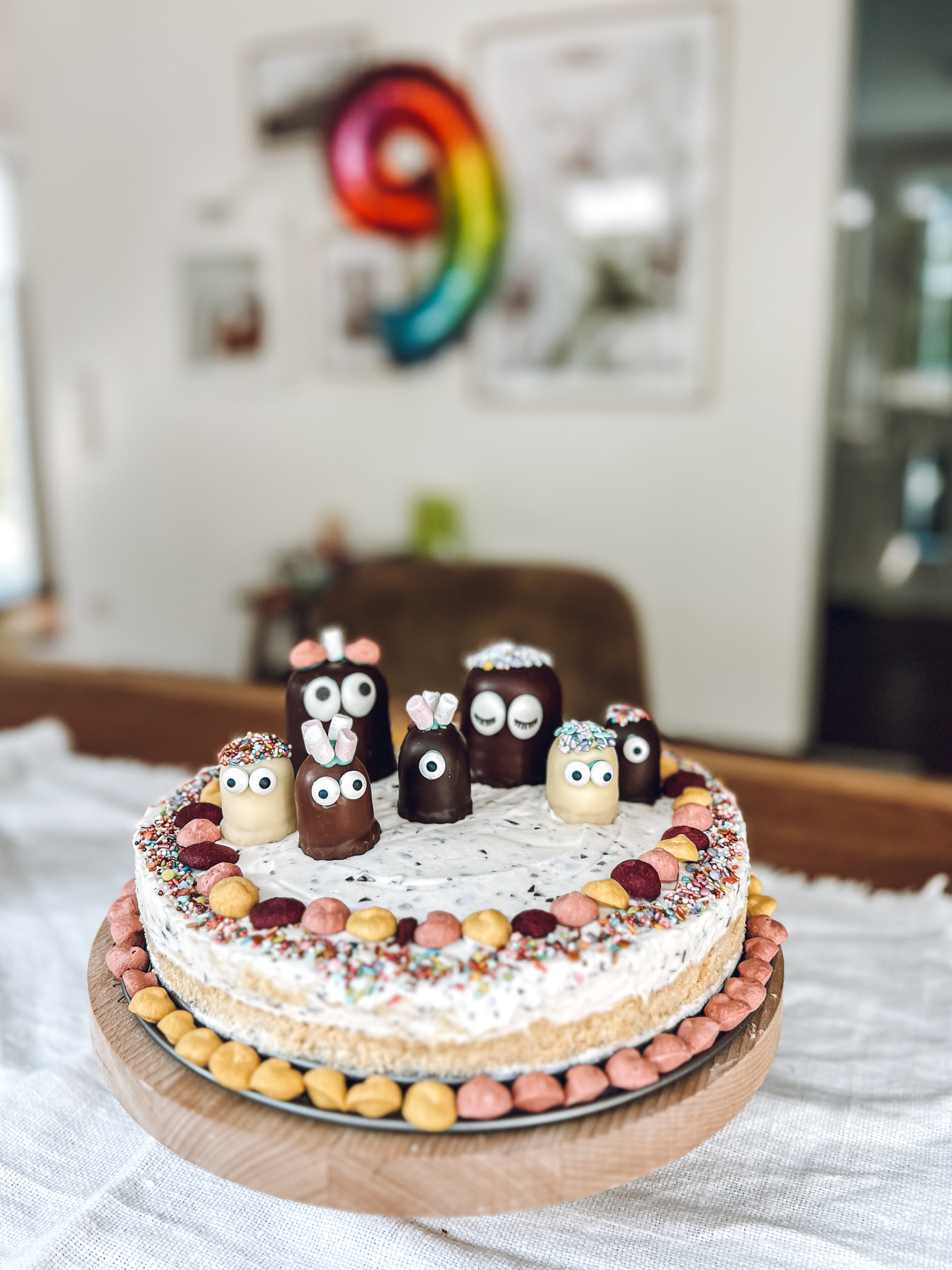 Bunte Geburtstagstorte mit dekorativen Figuren aus Schokolade und bunten Streuseln, auf einem Holzuntersetzer präsentiert.