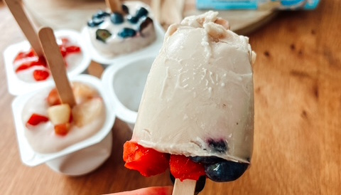 Fruchtiges Jogurt-Eis am Stiel mit Erdbeeren und Heidelbeeren, umgeben von frischen Beeren und Joghurtbechern auf einem Holztisch.