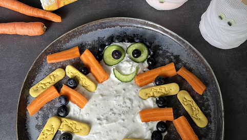 Bunte Spinne aus Quark, Karotten, Gurken, blauen Beeren und Alete Kinder-Crackern auf einem schwarzen Hintergrund, dekoriert mit Spuklichtern.