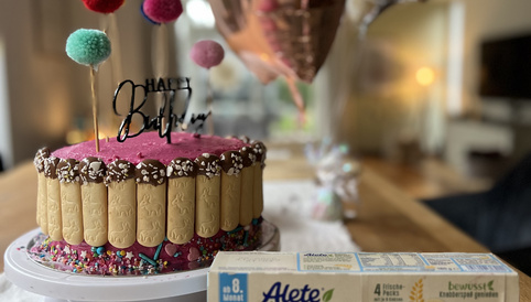 Bunte Geburtstagstorte mit Alete-Kinderkeksen und Dekoration aus Pompons, neben einer Packung Alete Kinderkekse auf einem Holztisch.