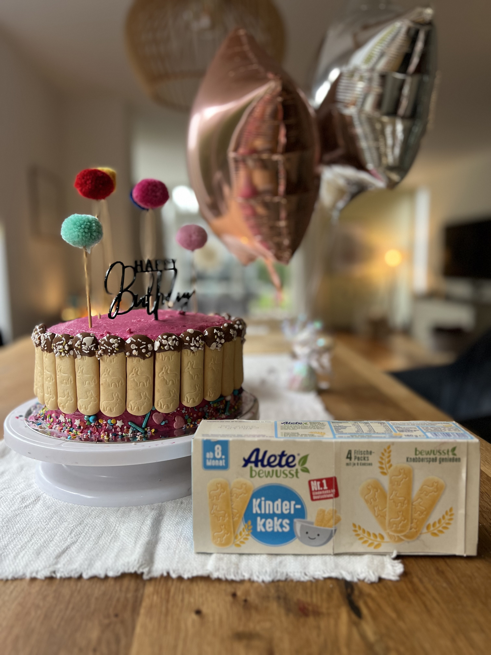 Bunte Geburtstagstorte mit Alete-Kinderkeksen und Dekoration aus Pompons, neben einer Packung Alete Kinderkekse auf einem Holztisch.